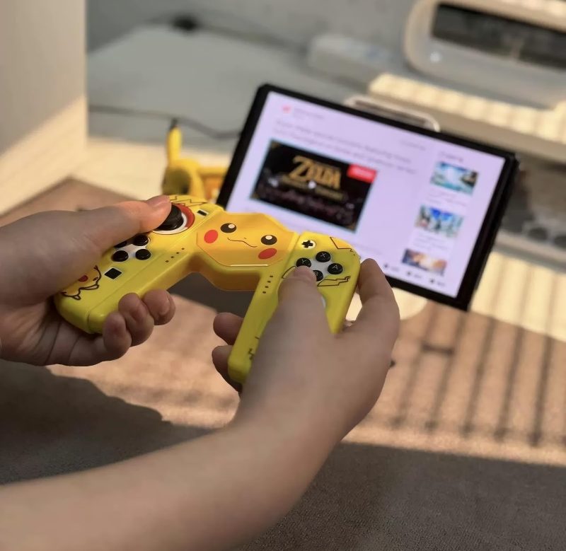 Manos sosteniendo un cargador de joycon inalámbrico amarillo con diseño de Pikachu frente a una tableta que muestra un video de juego de Zelda. El controlador presenta la cara de Pikachu y diseños de rayos, con botones y joysticks visibles